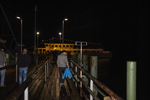 Mann auf einem Ufersteg mit beleuchtetem Schiff im Hintergrund.